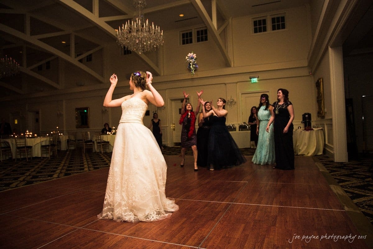 charlotte wedding photographers - ashley & matt A bride throws her bouquet over her shoulder to a group of women eagerly reaching for it on a dance floor. The venue is decorated with chandeliers and has a formal atmosphere.