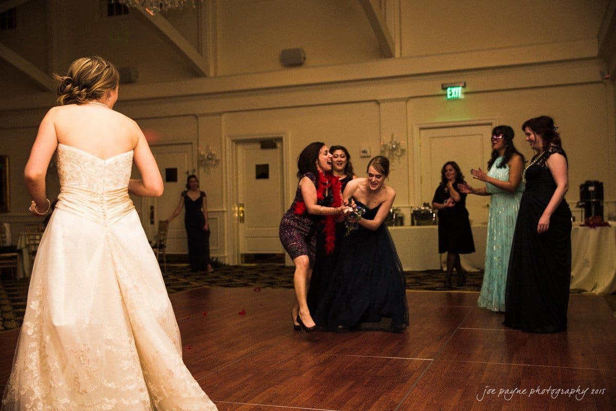 charlotte wedding photographers - ashley & matt A bride in a white dress watches as a group of women, dressed in gowns, excitedly gather on a dance floor. One woman in a navy dress holds a bouquet, smiling and leaning forward. The scene appears lively and celebratory.
