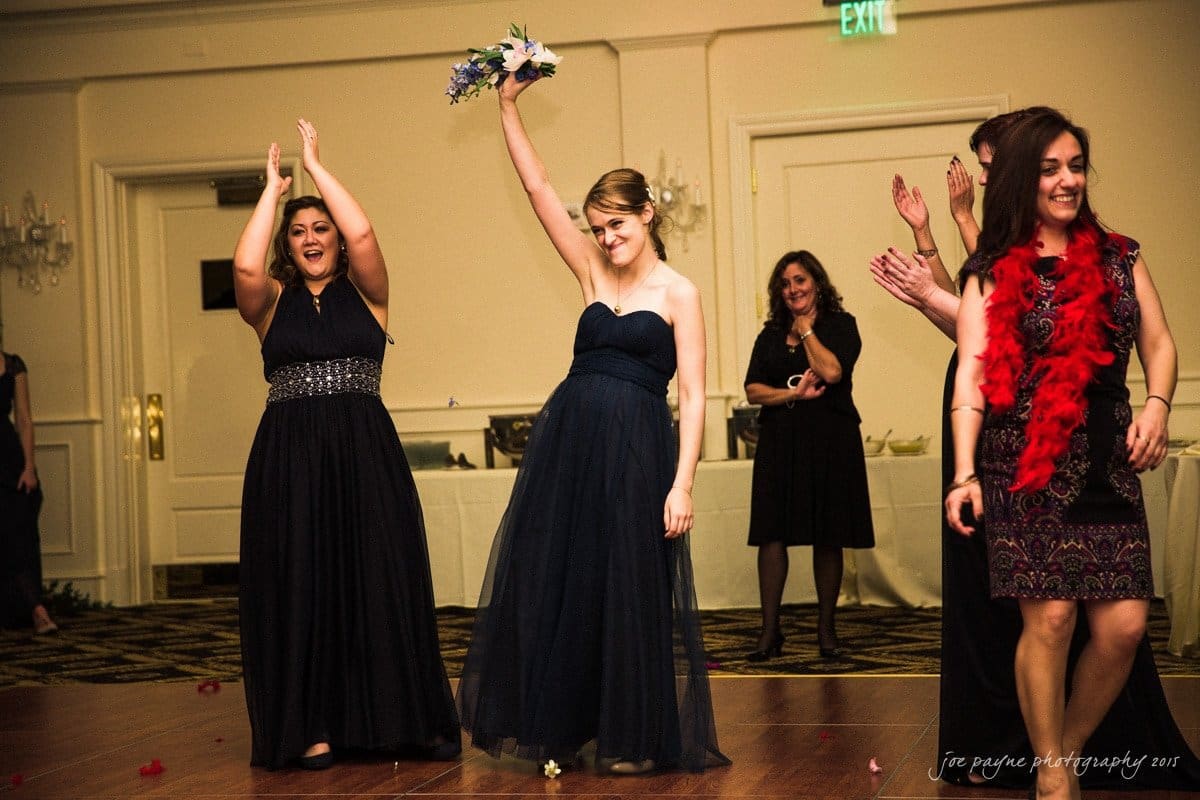charlotte wedding photographers - ashley & matt A group of women in formal attire celebrate a bouquet toss at a wedding reception. One woman holds the bouquet aloft, smiling, while others clap and cheer. The room is elegantly decorated with soft lighting.