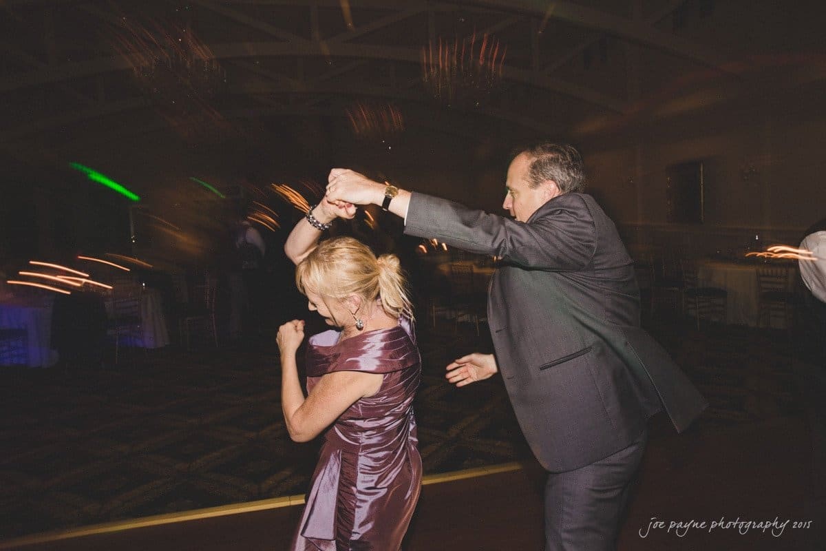 charlotte wedding photographers - ashley & matt A couple dances enthusiastically in a dimly lit room with blurred lights in the background. The woman wears a purple dress and the man is in a gray suit. They hold hands as they spin around the dance floor.