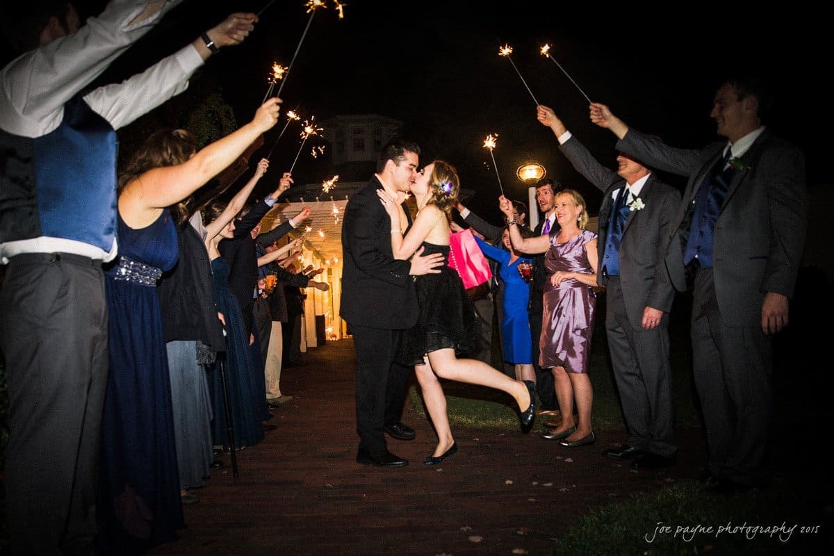 charlotte wedding photographers - ashley & matt A couple kisses under a canopy of sparklers held by cheering guests. The scene is set outdoors at night, with the couple in the center, surrounded by people forming a celebratory walkway.