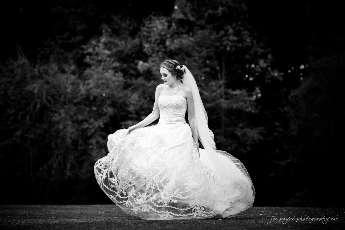 charlotte wedding photographers - ashley & matt A bride in a flowing gown holds her dress while standing outdoors. She has her hair styled with a veil, looking serenely to the side. The background is a blurred natural setting of trees, captured in black and white.