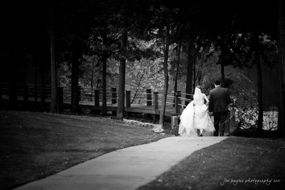 charlotte wedding photographers - ashley & matt A bride and groom walk hand in hand along a path surrounded by trees. The brides dress flows behind her, and the groom wears a suit. The scene is serene and intimate, with a bridge visible in the background.
