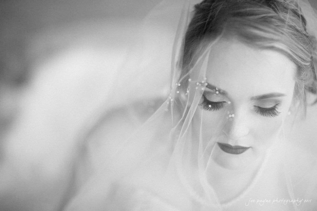 charlotte wedding photographers - ashley & matt A bride with closed eyes, wearing a veil adorned with small pearls. Her makeup features bold eyelashes and dark lipstick. The image is captured in black and white, creating a soft and serene atmosphere.