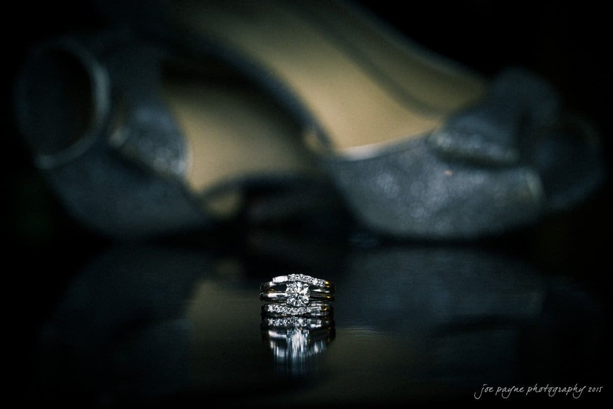 charlotte wedding photographers - ashley & matt A close-up of an elegant diamond ring with multiple bands, set against a blurred background of sparkling high-heeled shoes. The scene is dimly lit, highlighting the rings reflective shine.