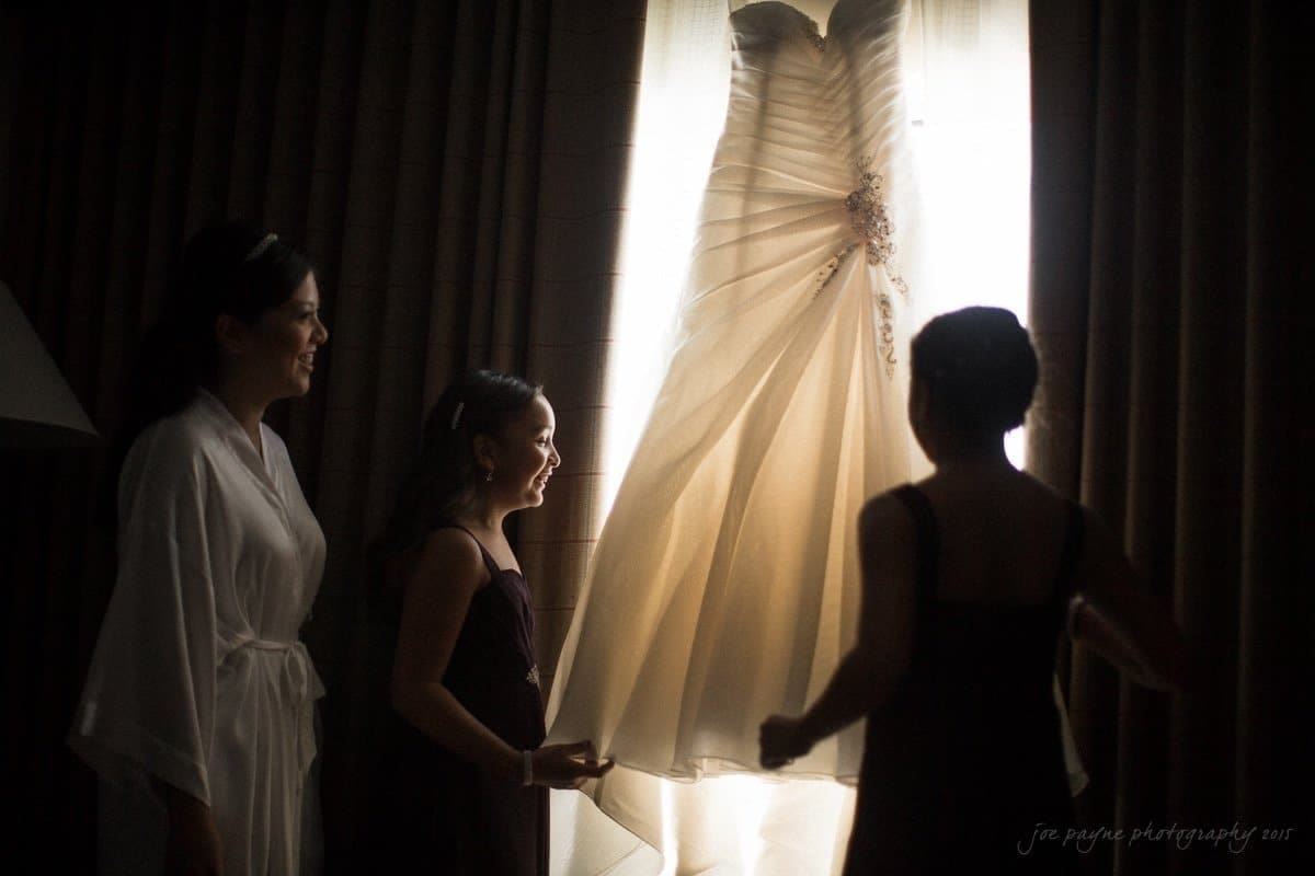 Two women and a girl admire a wedding dress hanging in front of a window. The dress glows softly in the natural light, highlighting its details. The atmosphere is warm and anticipatory.