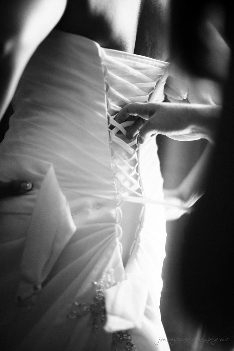 A black and white photo capturing a close-up of hands adjusting the lace-up back of a brides wedding dress, highlighting intricate details and the delicate fabric.