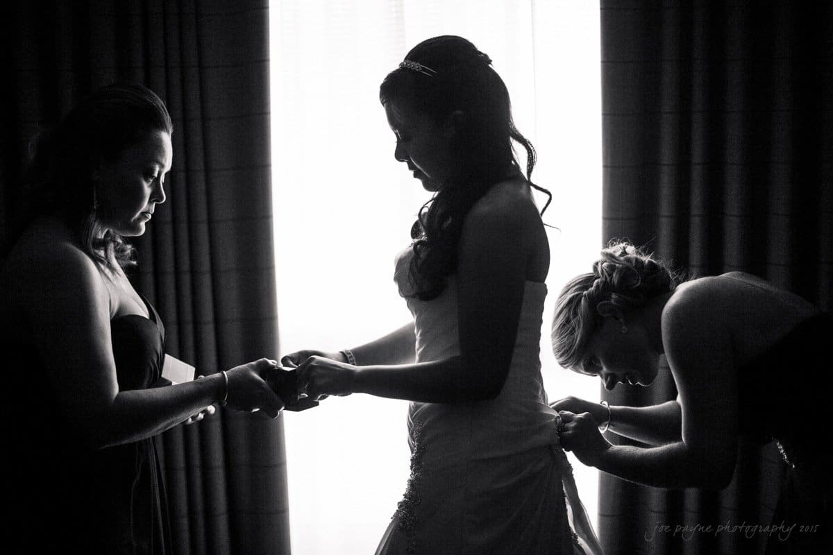 A silhouette of a bride being assisted by two bridesmaids. One adjusts her bracelet, while the other fixes her dress. Theyre standing in front of a softly lit window, creating a dramatic and elegant scene.