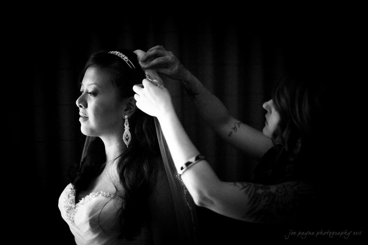 A bride stands in profile, illuminated by soft lighting. A woman adjusts her veil, showcasing detailed tattoos on her arm. The bride wears a strapless gown, a tiara, and elegant earrings. The background is dark, emphasizing the intimate moment.