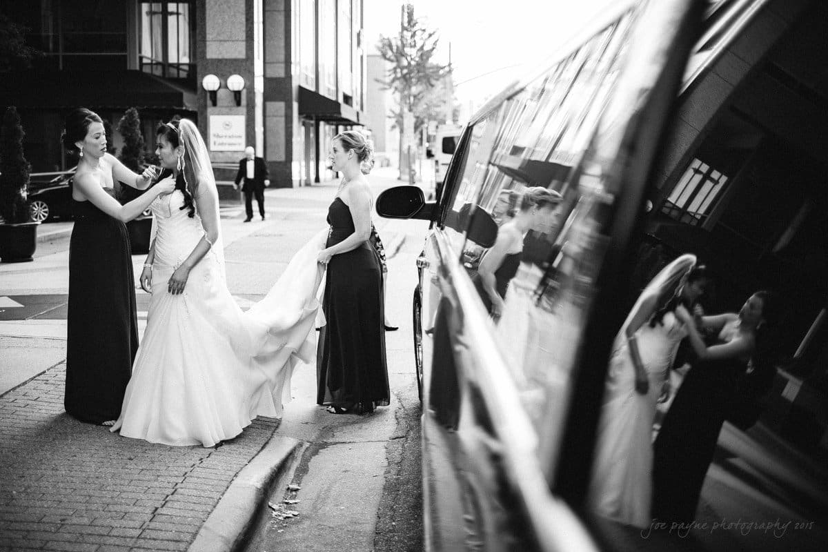 A bride in a wedding gown is being assisted by two bridesmaids on a city sidewalk. Reflections are visible in a car window in the foreground. The scene is captured in black and white.
