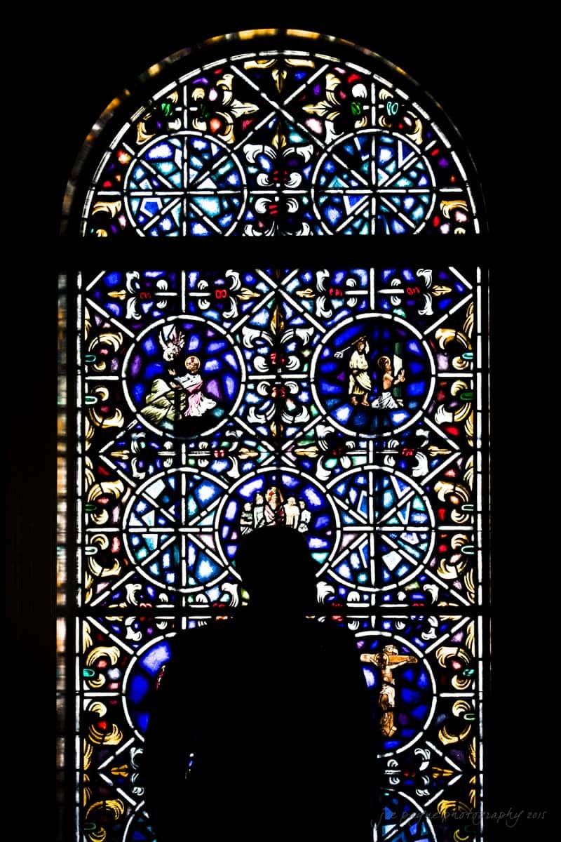 A person stands silhouetted in front of an ornate stained glass window. The window features intricate patterns and vibrant colors, creating a striking visual display against the darkened figure.