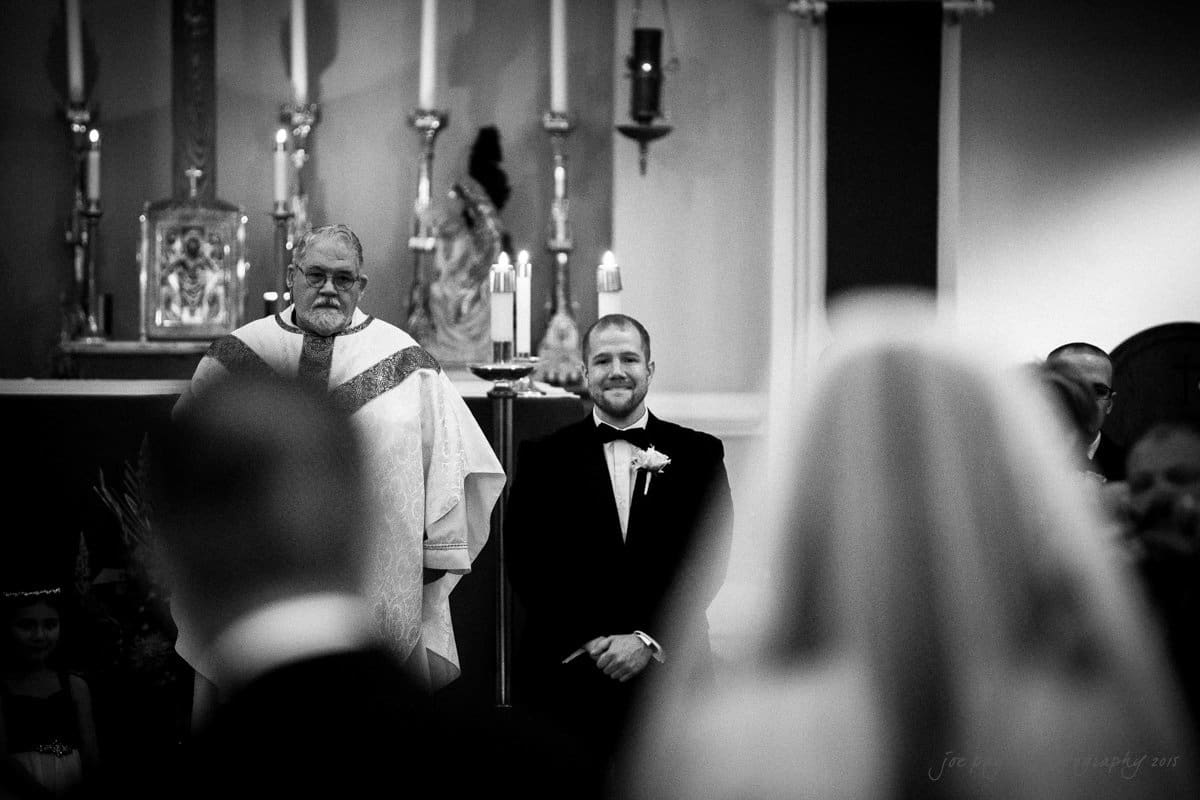 A black and white photo of a wedding ceremony. A man in a tuxedo stands smiling near a priest. Candles and religious decorations are in the background. A blurry bride is visible in the foreground.