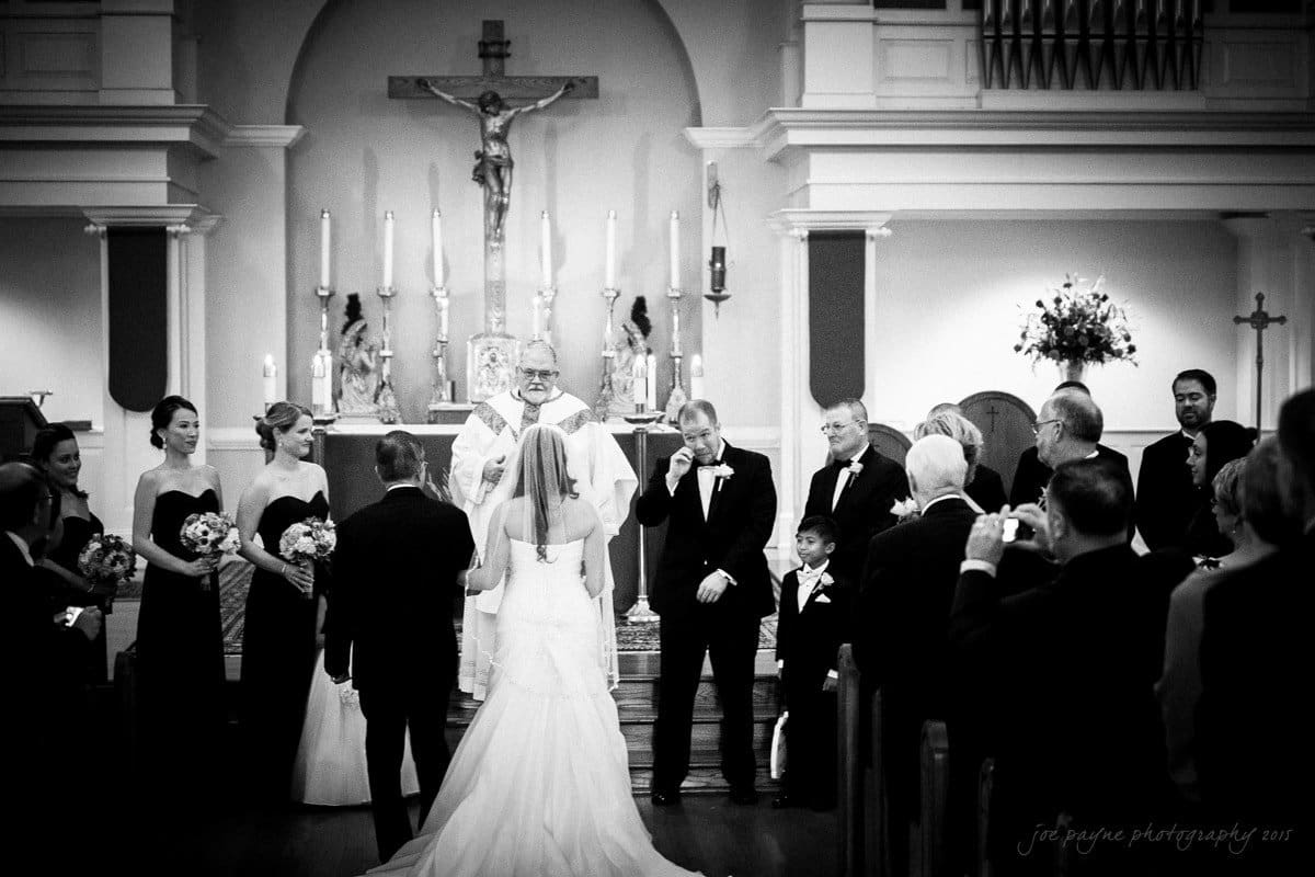 A black and white photo of a wedding ceremony in a church. The bride and groom stand before a priest at the altar. Bridesmaids in black dresses and groomsmen in suits surround them. Guests are seated, capturing the moment on their phones.