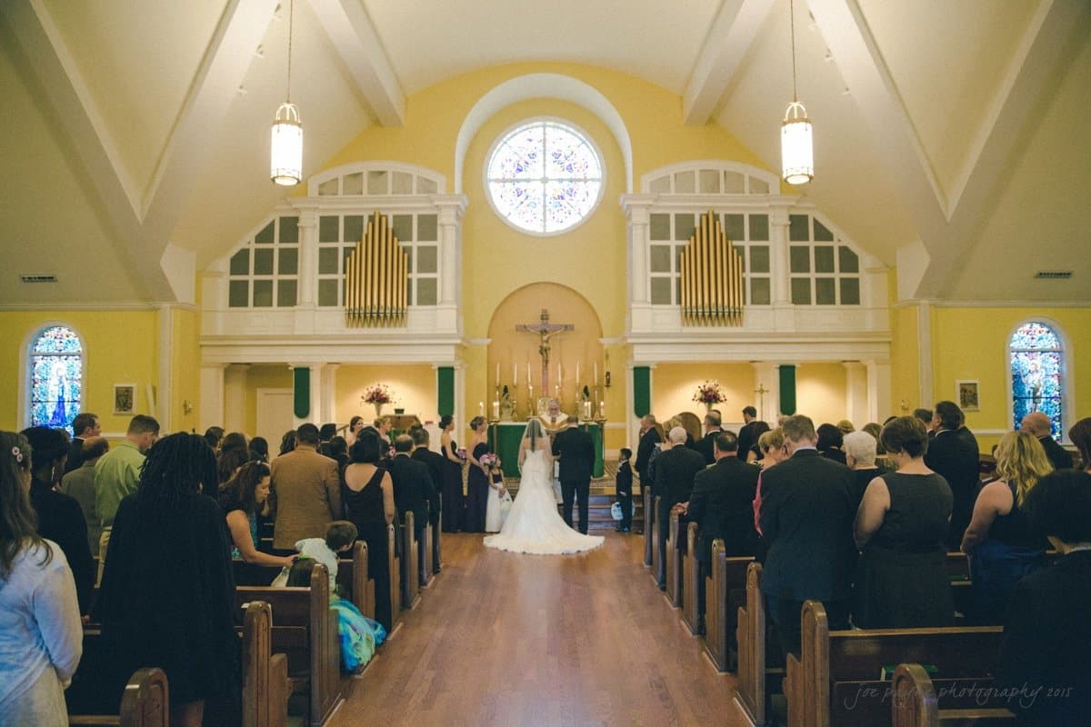 A wedding ceremony in progress inside a church. A bride and groom stand at the altar, surrounded by guests seated in pews. The church features stained glass windows, a large round window above the altar, and an organ.

.