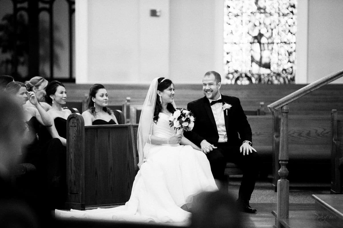 A bride and groom sit together in a church during their wedding ceremony, smiling and holding hands. Bridesmaids in dark dresses are seated nearby, looking towards the couple. A stained glass window is visible in the background. The image is in black and white.