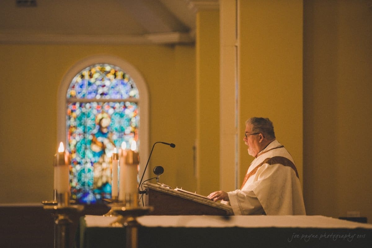 A priest stands at a pulpit in a church, reading from a book. Lit candles are in the foreground, and a stained glass window depicting a religious figure is visible in the background.