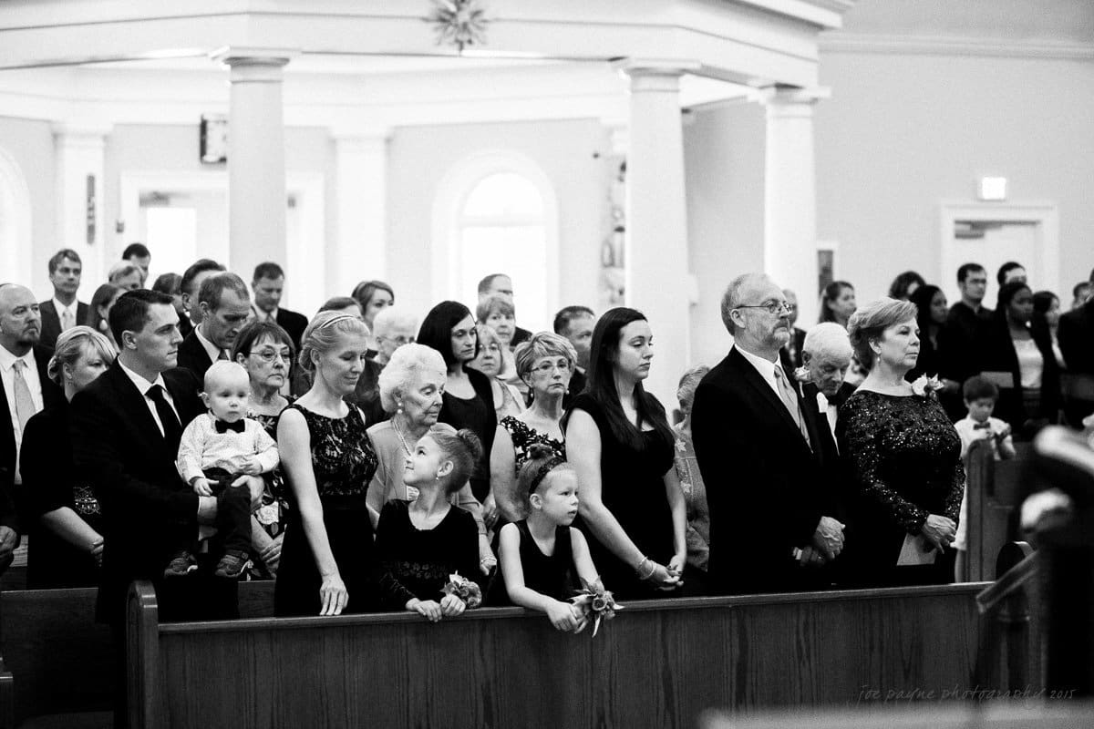 A black-and-white photo of a group of people in formal attire standing inside a church. Adults and children are lined up in several rows, some holding hands. The atmosphere appears solemn and attentive.