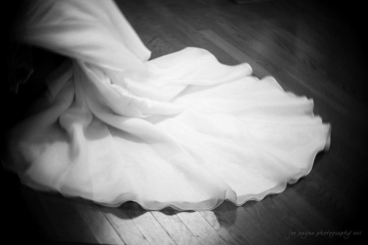A flowing white gown swirls elegantly on a polished wooden floor, creating a sense of motion and grace. The monochrome effect highlights the texture and folds of the fabric.