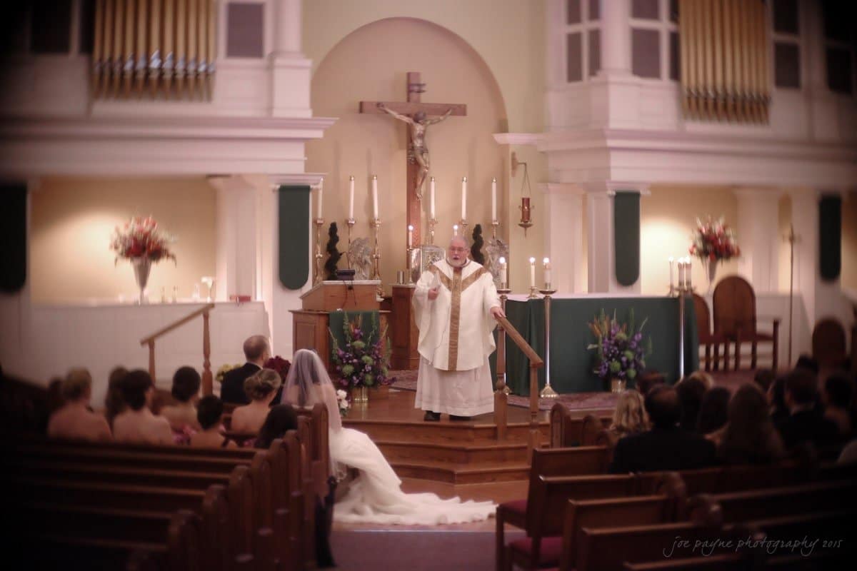 A priest officiates a wedding ceremony in a church. The bride and groom are seated in the front pew, with guests attending. The altar is decorated with candles, flowers, and a crucifix in the background.