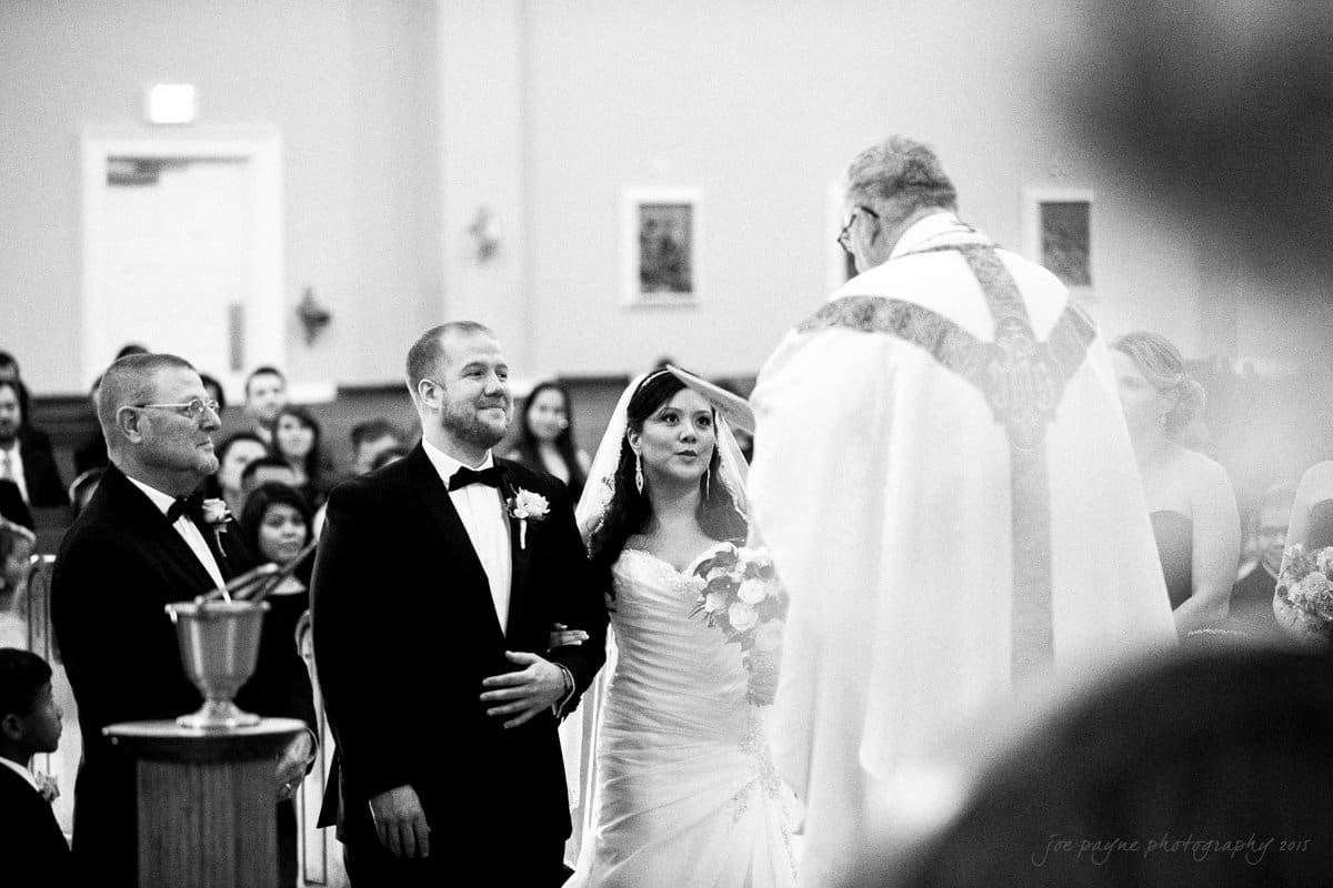 A bride and groom stand before a priest during a wedding ceremony in a church. The groom is in a tuxedo, and the bride wears a white gown, holding a bouquet. The priest blesses them as guests watch from the pews.