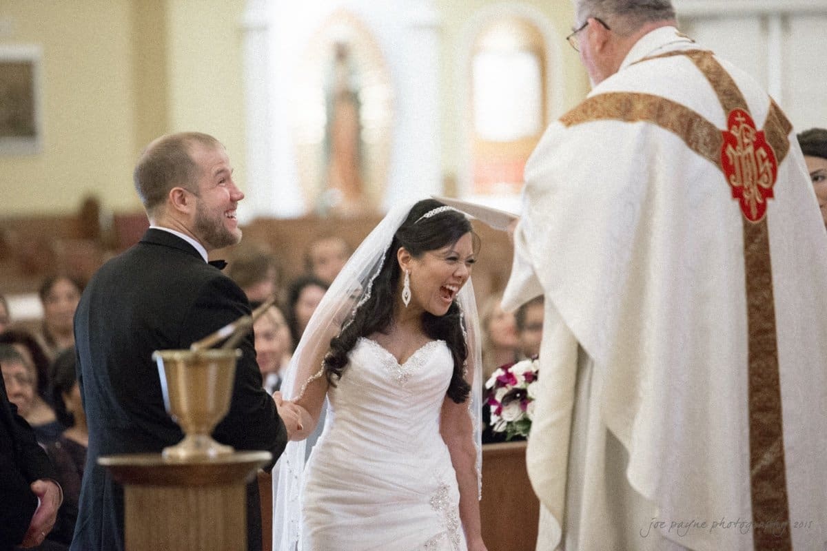 A bride and groom stand at the altar during their wedding ceremony, facing a priest. The bride is laughing joyfully while wearing a white gown and veil, and the groom is in a black suit. The setting is a church with seated guests in the background.