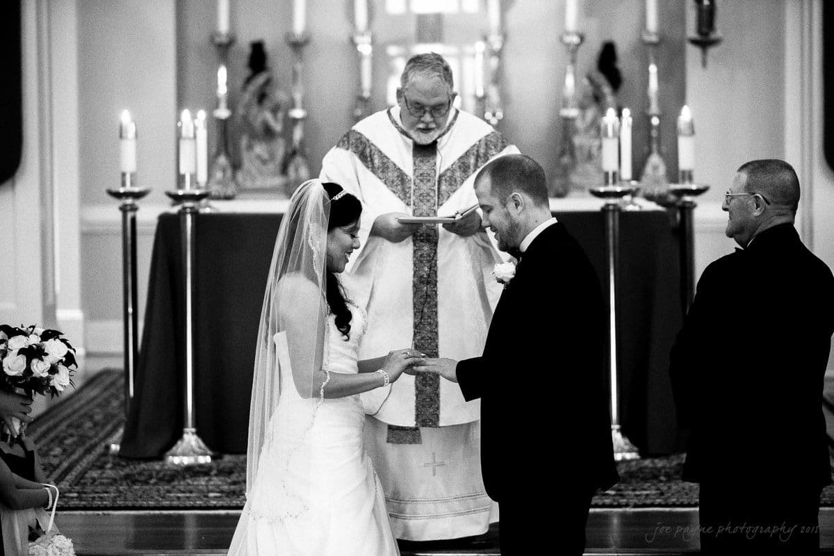 A bride and groom exchange rings during a wedding ceremony in a church. The officiant stands behind them in front of an altar adorned with candles. Bridesmaids hold flowers on the left, and a groomsman stands on the right.