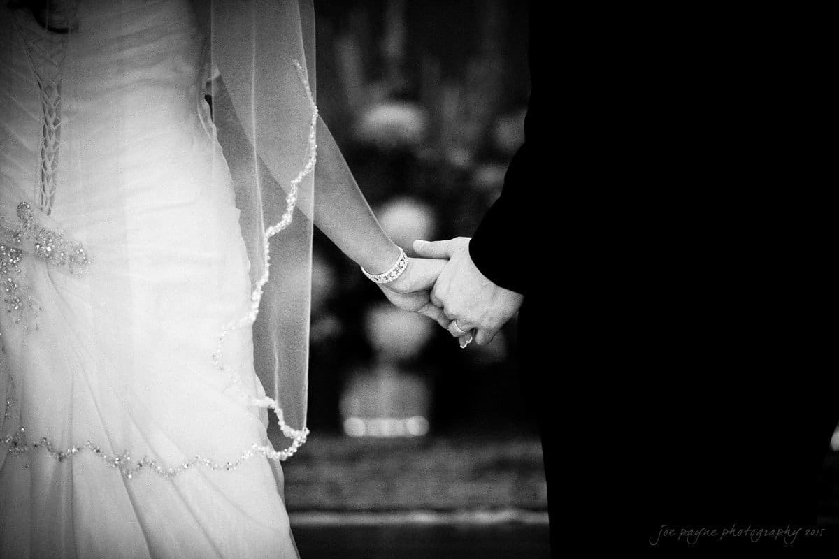 A black and white photo showing a bride and groom holding hands. The bride wears a gown with lace detailing and a bracelet, while the groom is in a dark suit. Their clasped hands are the focal point in the foreground.