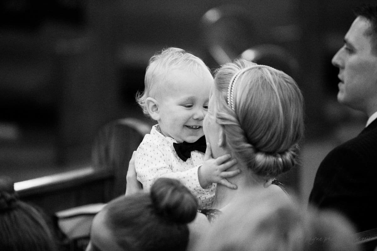 A black and white photo of a smiling young child wearing a bow tie, held by an adult with neatly styled hair. The scene appears to be indoors, possibly during a formal event, with other individuals partially visible in the background.