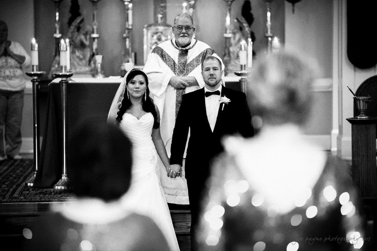 A bride and groom stand hand in hand at the altar during a wedding ceremony. A priest smiles behind them. They face the guests, including two blurred figures in the foreground. Candles and religious artifacts are visible in the background.