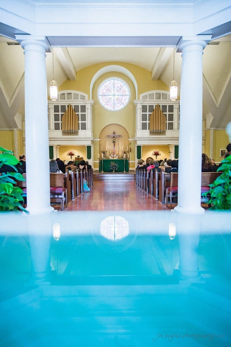 Interior of a church with white columns and a pool of water in the foreground. The church is filled with people seated, and a stained glass window and organ pipes are visible at the front.