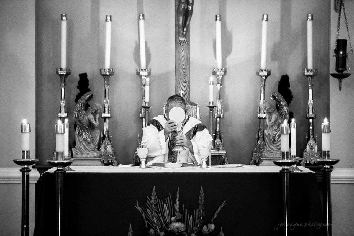 A priest stands at an altar, holding up a white host during a religious ceremony. The background features tall candles and statues. The setting is ornate and sacred, with a focus on the ritual being performed.