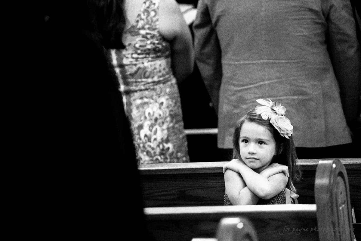A black and white image of a young girl sitting alone in a pew at church. She has a large flower headband and is looking contemplatively ahead, with her arms crossed on the bench back. Blurred adults are standing in the background.