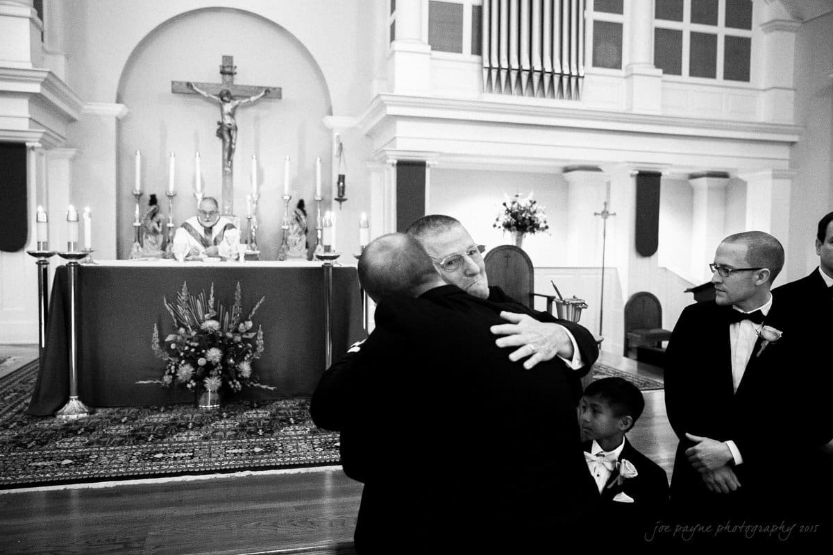 A man in a suit hugs another man during a church ceremony. A young boy stands nearby, dressed formally. The altar, decorated with candles and flowers, is visible in the background, along with a priest seated on a high chair.