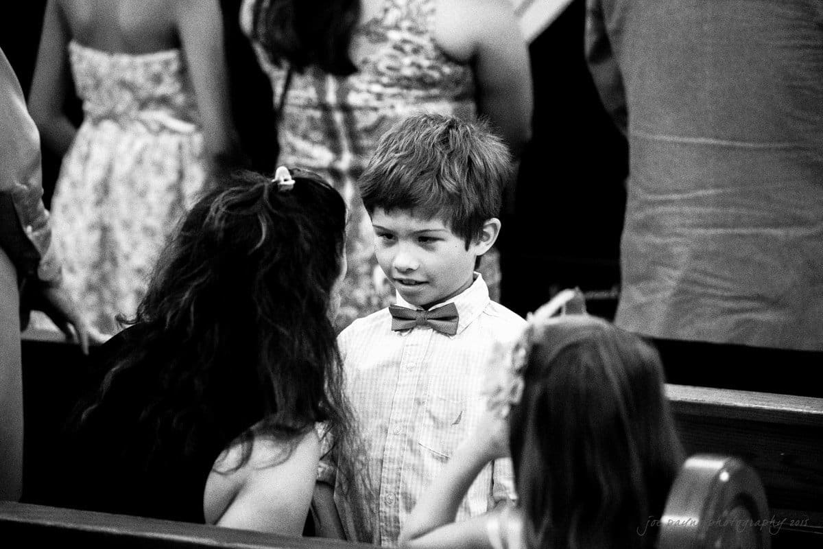 A black-and-white photo shows a young boy with a bow tie sitting in church pews, surrounded by others. He is facing a woman who is speaking to him. The scene captures an intimate moment during a gathering.