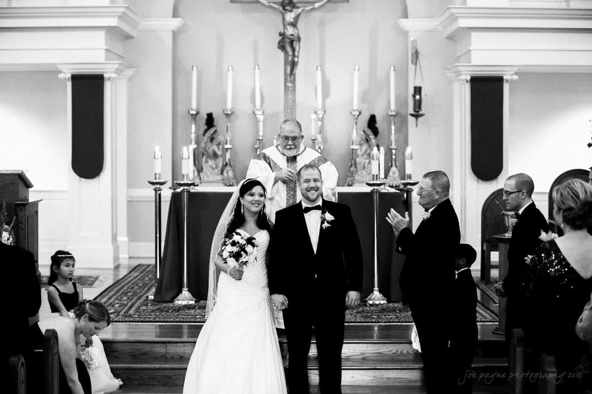A bride and groom stand at the altar during a church wedding ceremony. The bride holds a bouquet, and a clergyman stands behind them with a smile. Guests are seated nearby, and the venue is ornate with lit candles.