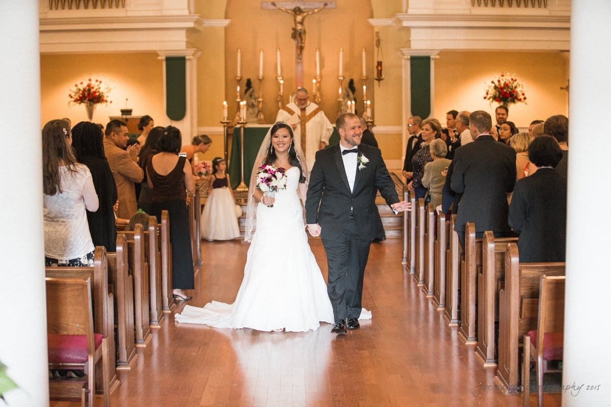 A bride and groom walk down the aisle hand in hand in a church. The bride wears a white gown and holds a bouquet, while the groom is in a black tuxedo. Guests stand in the pews, and a priest is visible at the altar in the background.