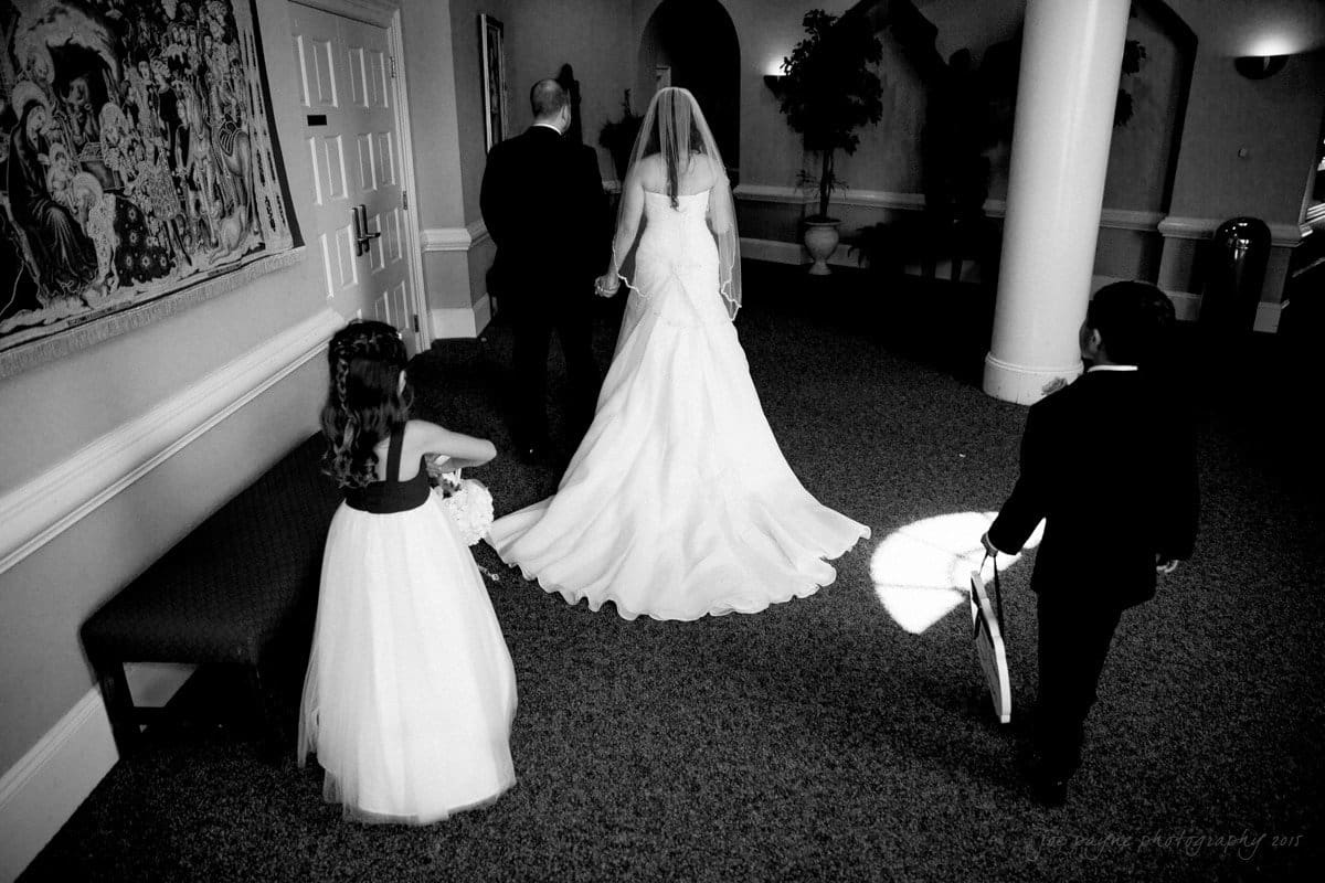 A bride and groom walk hand in hand down a hallway. The bride wears a long white gown with a veil. Ahead of them, a young girl in a white dress and a boy in a dark suit hold flower baskets, leading the way.