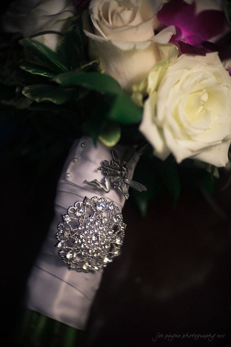 A bridal bouquet featuring white roses, purple flowers, and green leaves. The stems are wrapped in satin fabric adorned with a sparkling brooch.