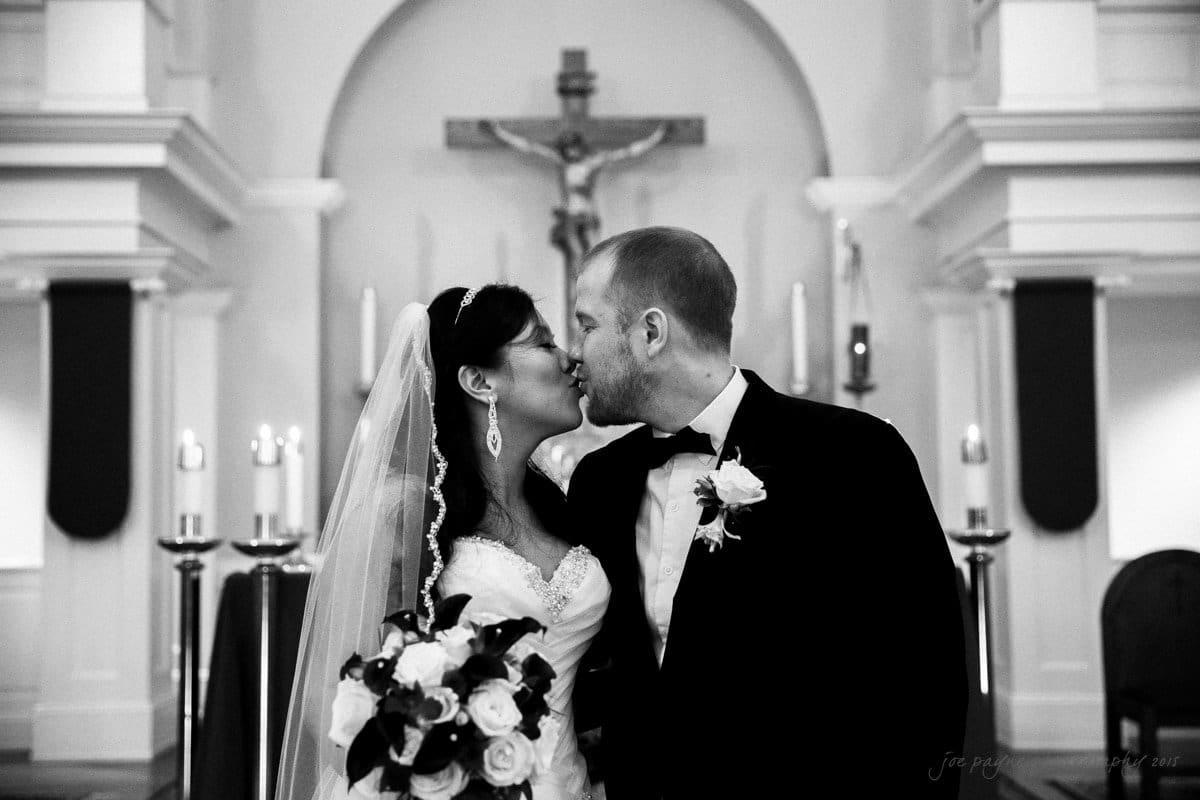 A bride and groom kiss in a church. The bride holds a bouquet and wears a veil and earrings. The groom wears a suit with a boutonniere. Candles and a cross are in the background. The image is in black and white.