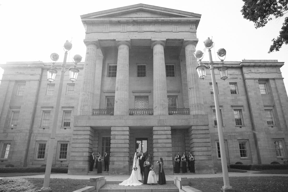 A black and white photo of a wedding ceremony taking place on the steps of a grand historic building with large columns. The couple is surrounded by their wedding party and guests, under tall street lamps.