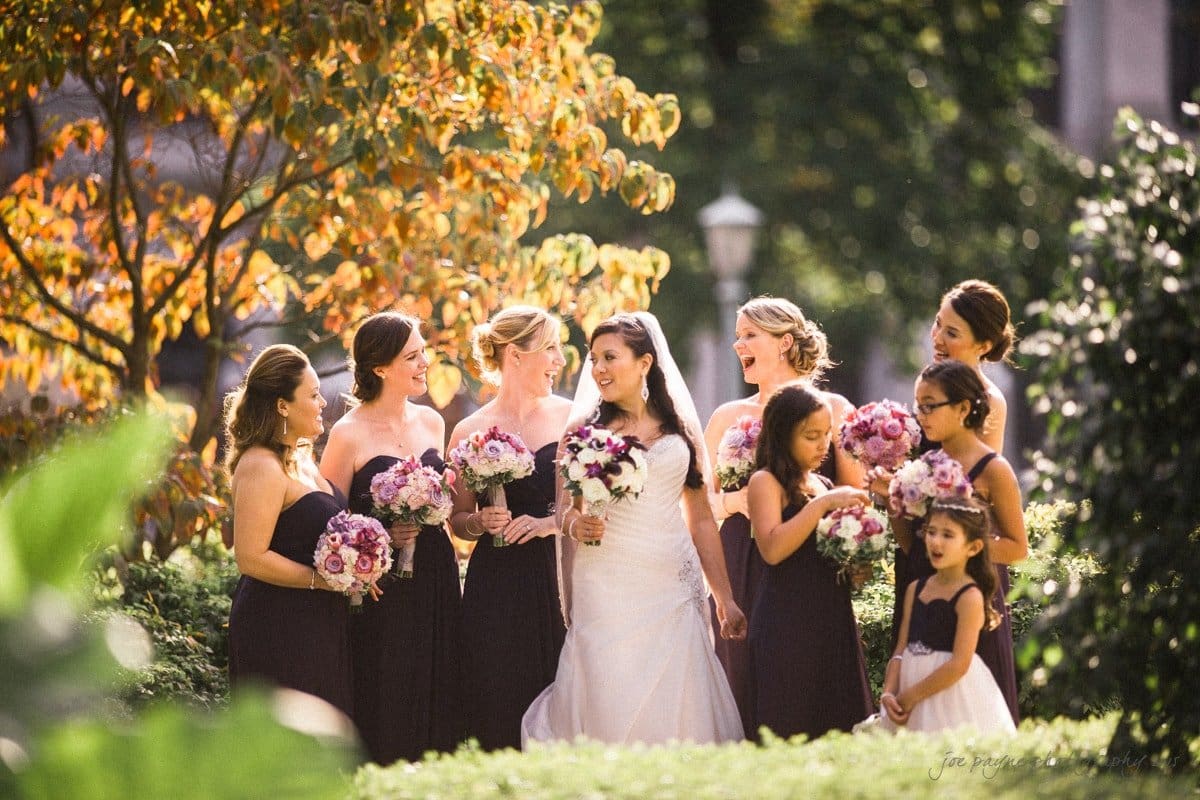 A bride in a white gown stands in a garden, surrounded by bridesmaids in dark dresses holding bouquets. Flower girls in similar dresses stand nearby. The scene is sunny, with colorful autumn leaves in the background.