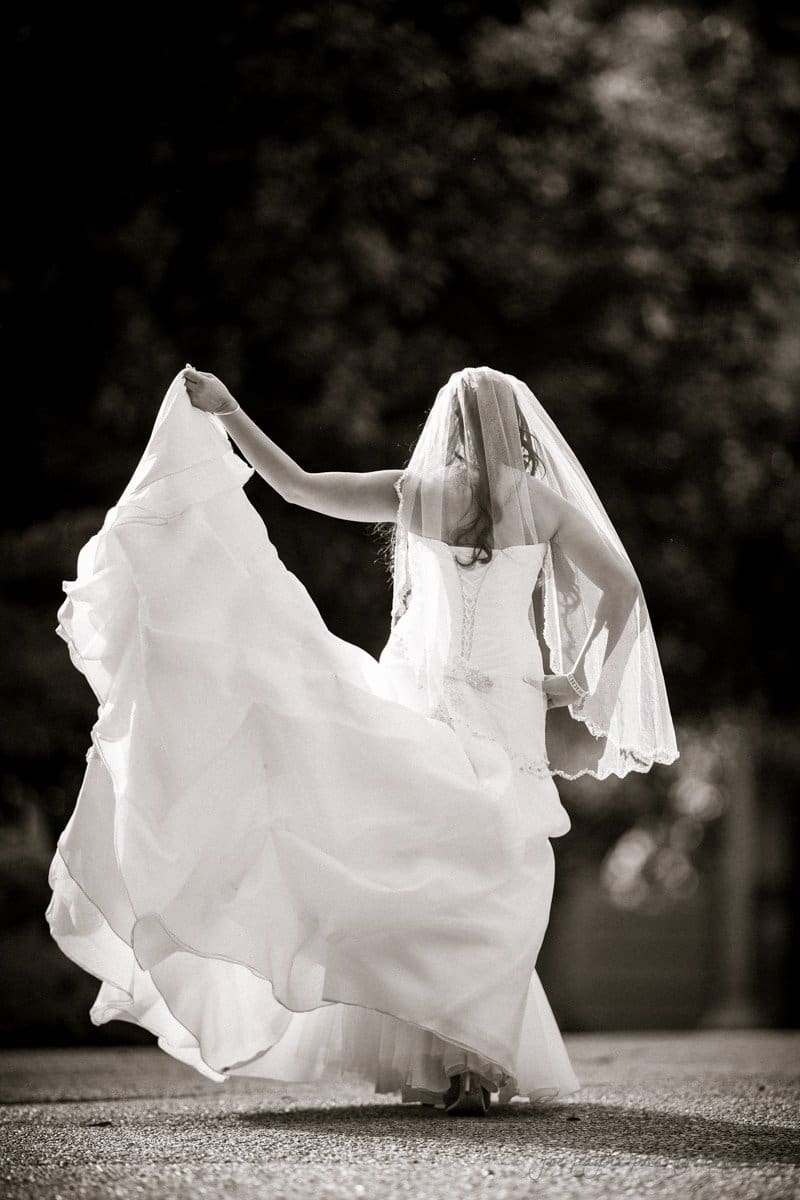 A bride in a flowing wedding gown and veil twirls outdoors, her dress billowing around her. Sunlight filters through the trees, creating a serene, joyful atmosphere. The image is in black and white.