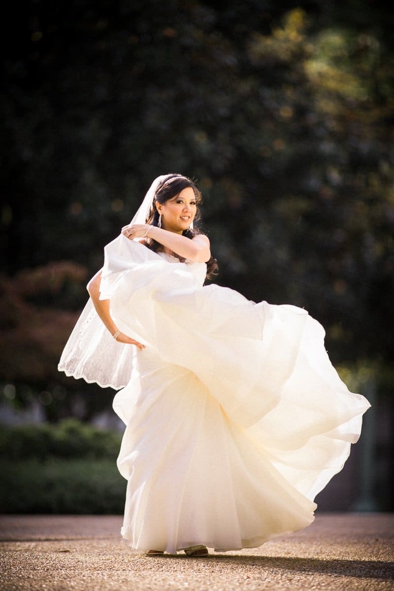 A bride in a flowing white gown twirls joyfully outdoors. Her veil catches the light as she spins, creating a graceful, dynamic motion. The background is softly blurred greenery, adding a romantic touch to the scene.