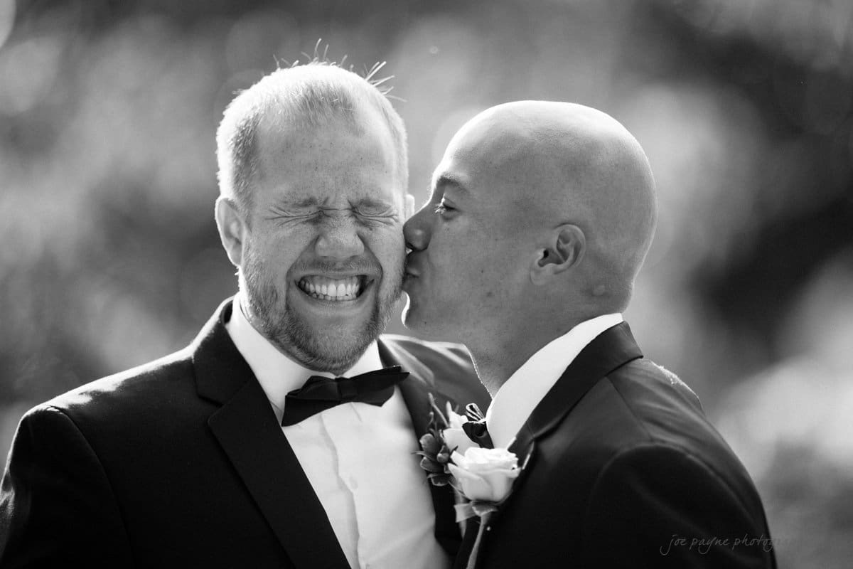 Two men in suits share a joyful moment; one has his eyes closed, smiling widely, while the other gives him a kiss on the cheek. The background is softly blurred.