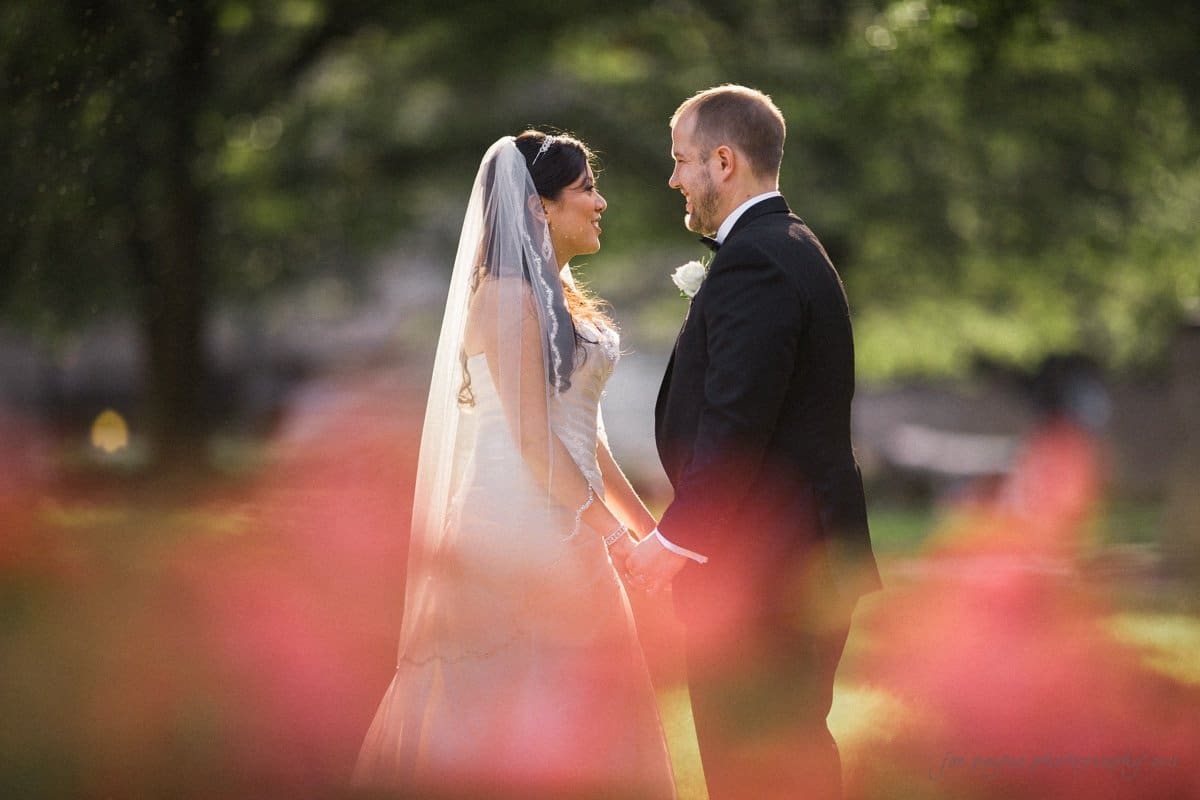 A bride and groom stand together outdoors, holding hands and facing each other. The bride is in a white dress and veil, and the groom is in a black suit. They are surrounded by greenery and blurred pink flowers in the foreground.