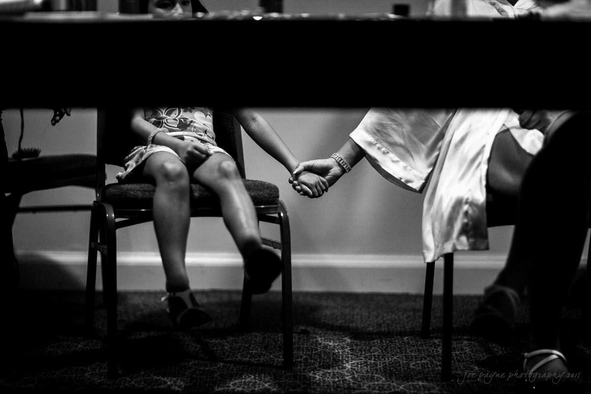 In a black and white photo, a child and an adult are seated next to each other, holding hands under a table. Their faces are not visible, creating a sense of intimacy and connection. The scene is set indoors with a carpeted floor.