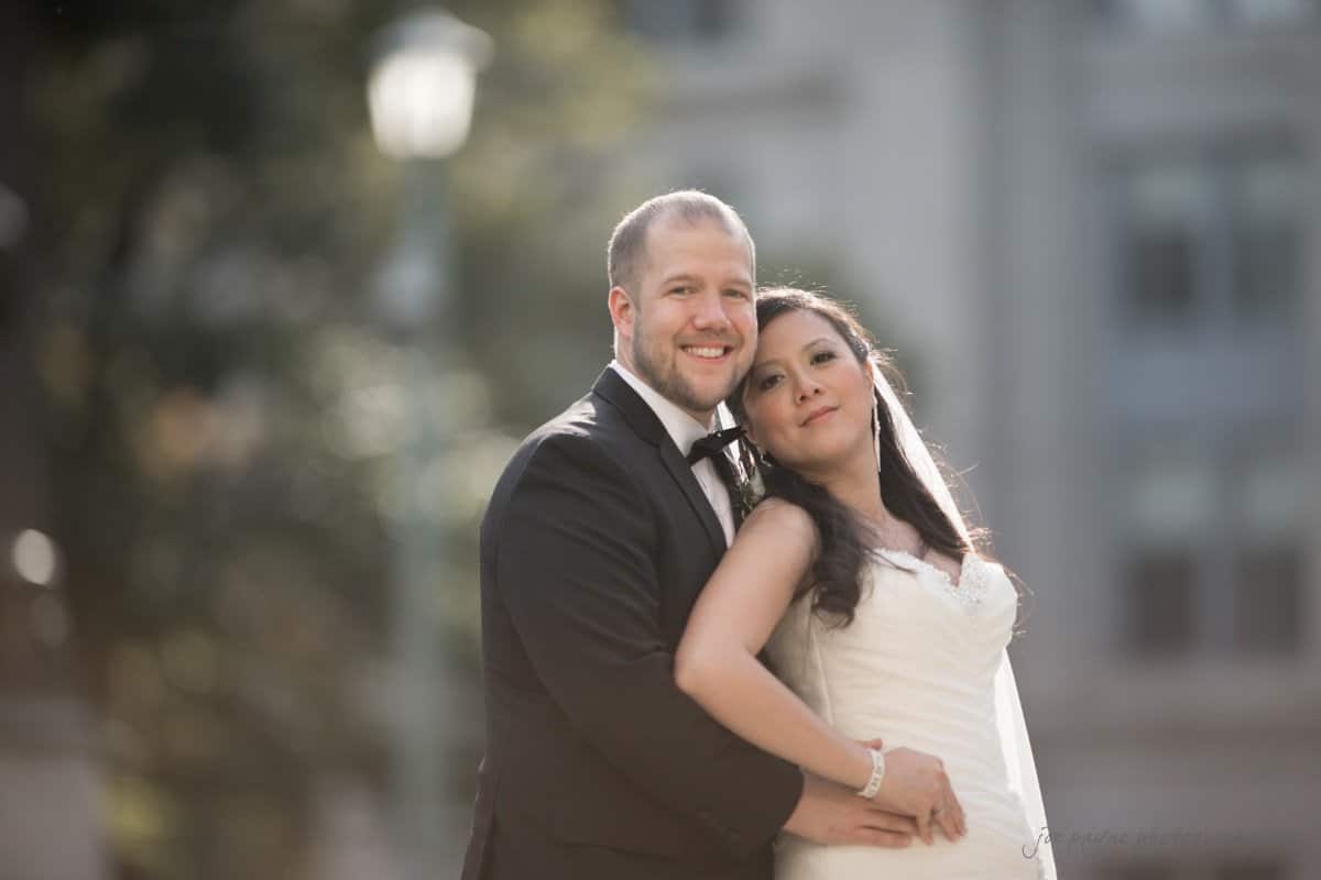 A bride and groom in wedding attire pose lovingly outdoors. She wears a white gown and veil, while he wears a black tuxedo. They are standing closely, with a blurred background of greenery and buildings.