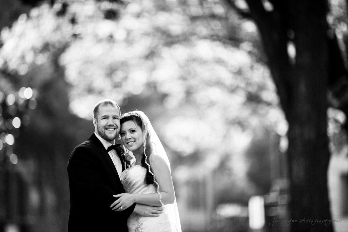 A bride and groom stand closely together outdoors, smiling and holding hands. The bride wears a veil and a white dress, while the groom is in a dark suit with a bow tie. The background is softly blurred, suggesting a tree-lined path.