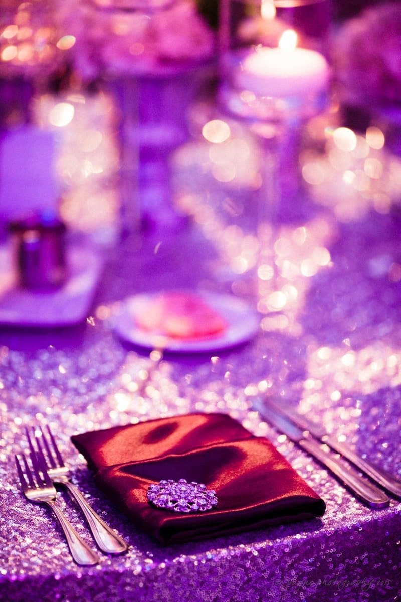 A dimly lit, elegant table setting featuring a shiny purple tablecloth, a place setting with a folded maroon napkin and a sparkling brooch, surrounded by cutlery. The background has blurred twinkling lights and a lit candle, creating a romantic ambiance.