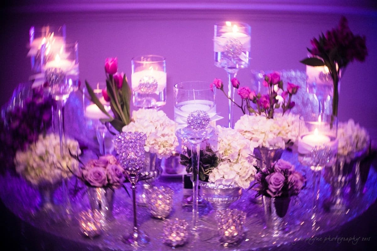 A dimly lit, sparkling table centerpiece with pink tulips, white flowers, candles in glass holders, and crystal ornaments, all set against a purple background.